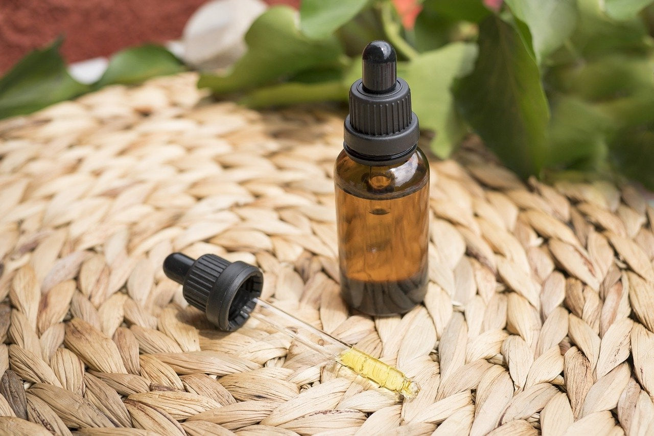 An amber glass dropper bottle with a black cap stands on a woven straw mat, with the cap placed beside it and a small yellow vial lying nearby. Green leaves are blurred in the background.
