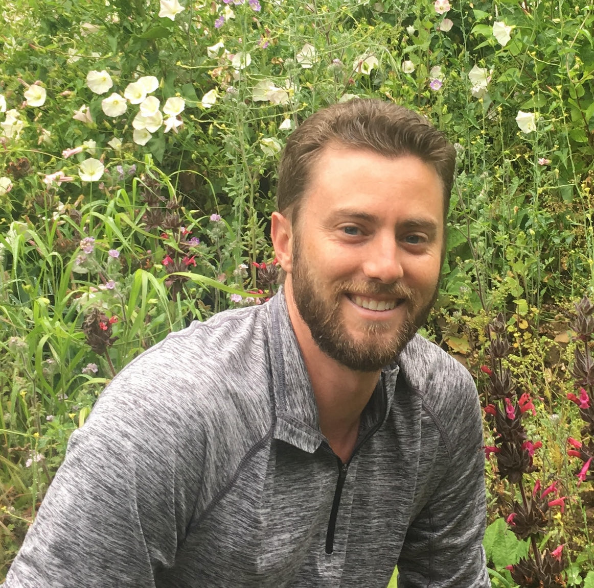 Man with a beard sitting in a garden with flowers and plants